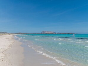 Una spiaggia con acque cristalline turchesi, sabbia bianca e montagne in lontananza sotto un cielo azzurro intenso.