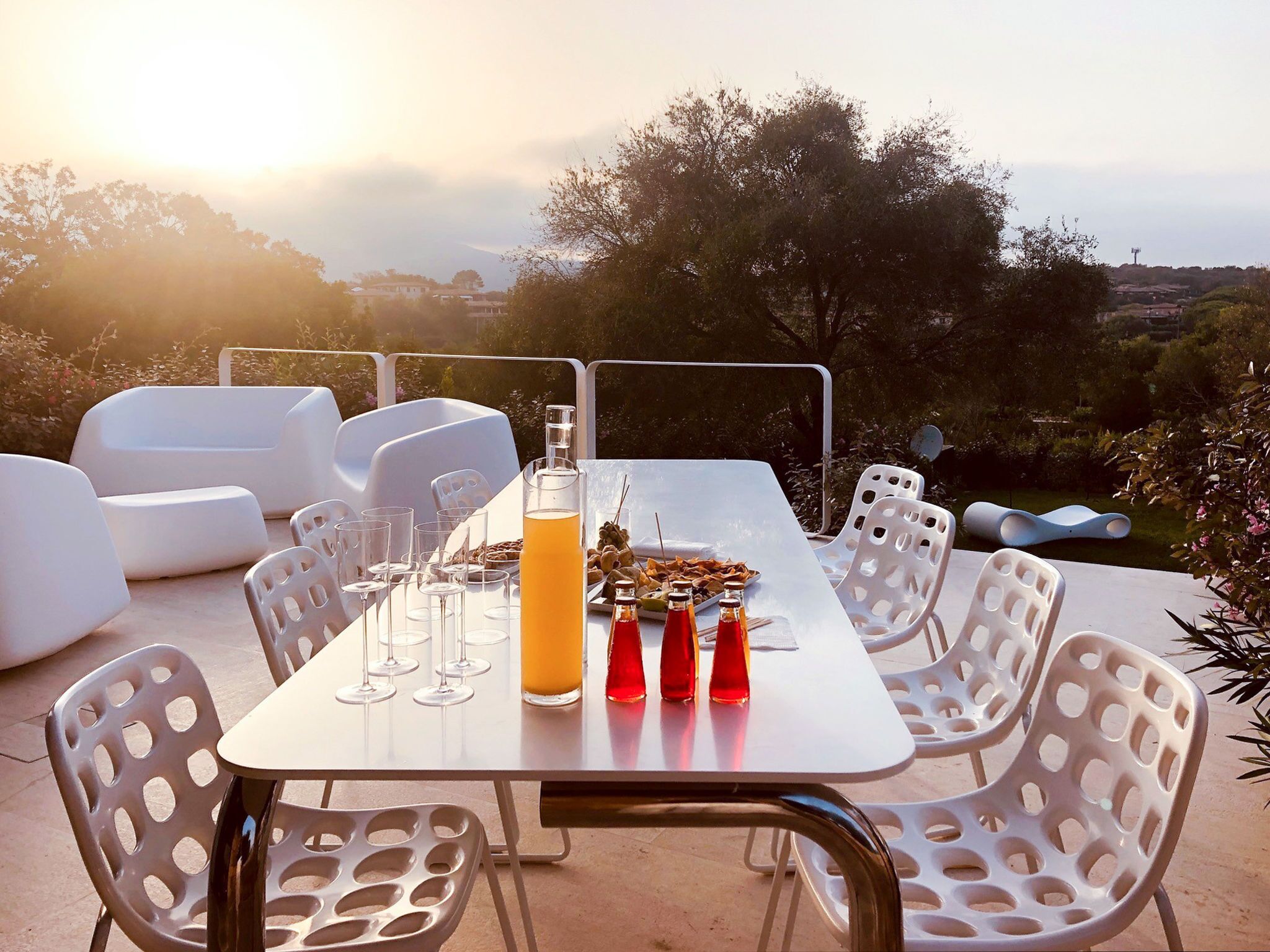 An outdoor dining area with a table set for four, surrounded by white plastic chairs. There are wine glasses, a bottle of orange juice, and two bottles of red drink on the table.