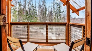 A wooden cabin with a large window overlooking a forest. Inside, there are two chairs and a table.