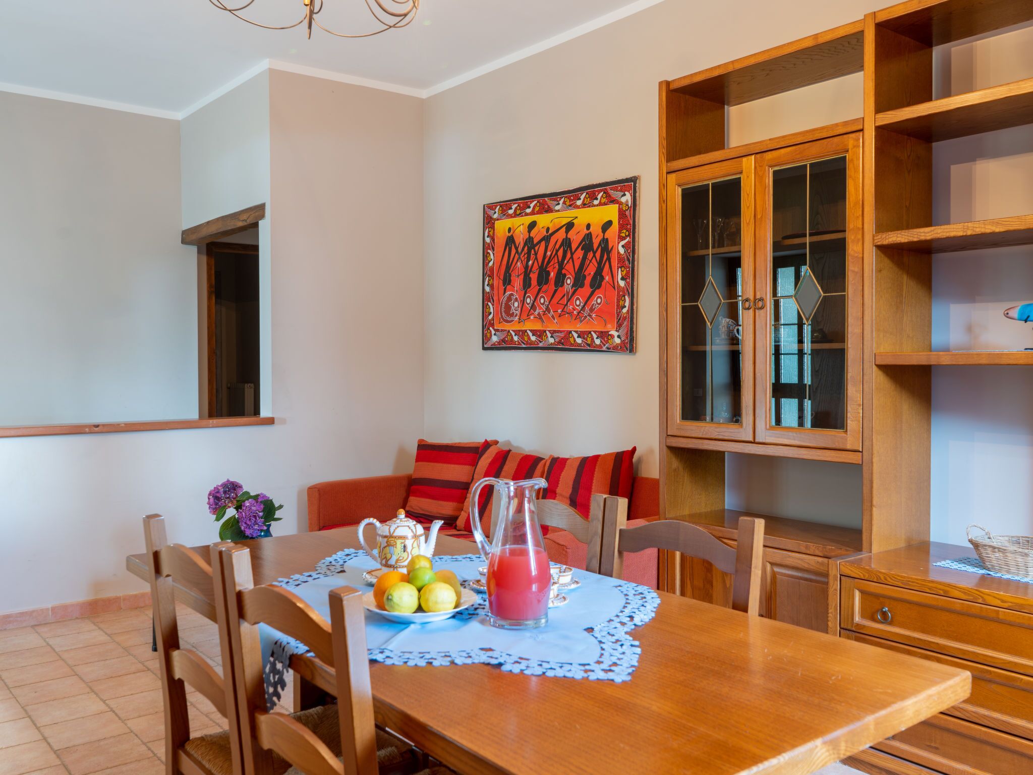 A dining area with a wooden table, chairs, a vase with flowers, a pitcher of juice, and a plate of fruit.