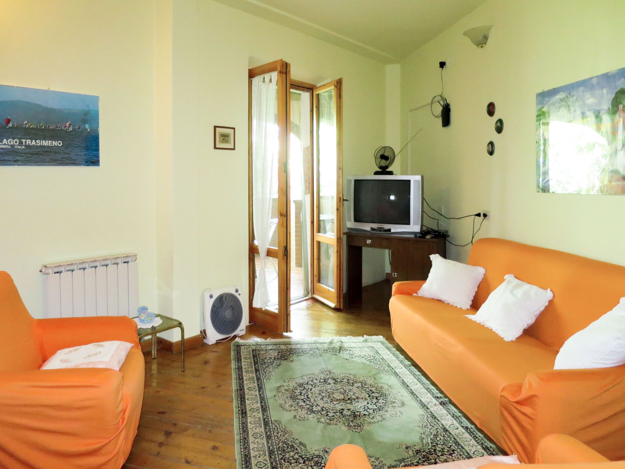 Een woonkamer met een oranje bank, een televisie op een houten dressoir, een ventilator en een groot raam met gordijnen.