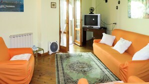 A living room with an orange sofa, a television on a wooden stand, a fan, and a large window with curtains. - Vacanza L'Olivo by Interhome (Lago Trasimeno)