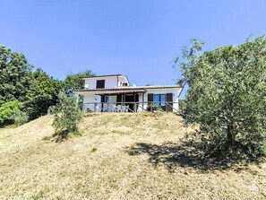 Una casa a due piani con un balcone, circondata da alberi e un pendio erboso.