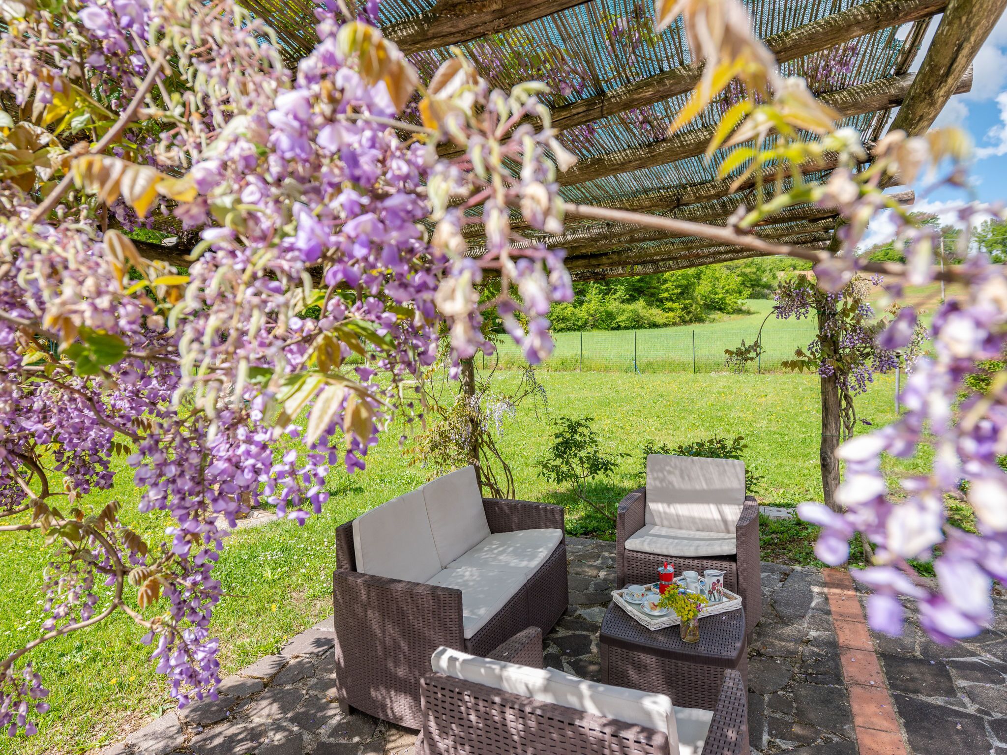 Un área de estar al aire libre con mobiliario de mimbre, una mesa con una bandeja de objetos y flores moradas en primer plano.