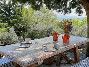 Mesa al aire libre con mantel de baldosas, dos teteras, tazas y un florero con flores.