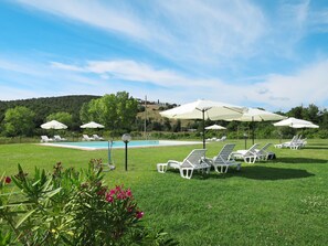 Zona junto a la piscina con sillas reclinables blancas y sombrillas, rodeada de vegetación y colinas al fondo.