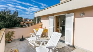 Um terraço com cadeiras brancas e uma mesa, um vaso de planta e vista para o bairro.