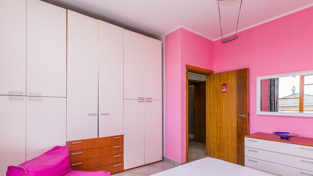 Quarto com parede rosa, guarda-roupa de madeira, cama branca e sofá rosa.