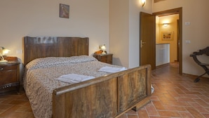 Ein Schlafzimmer mit einem Holzbett, zwei Nachttischen, einem Schminktisch und einem Gemälde an der Wand. Eine Tür führt in einen anderen Raum.