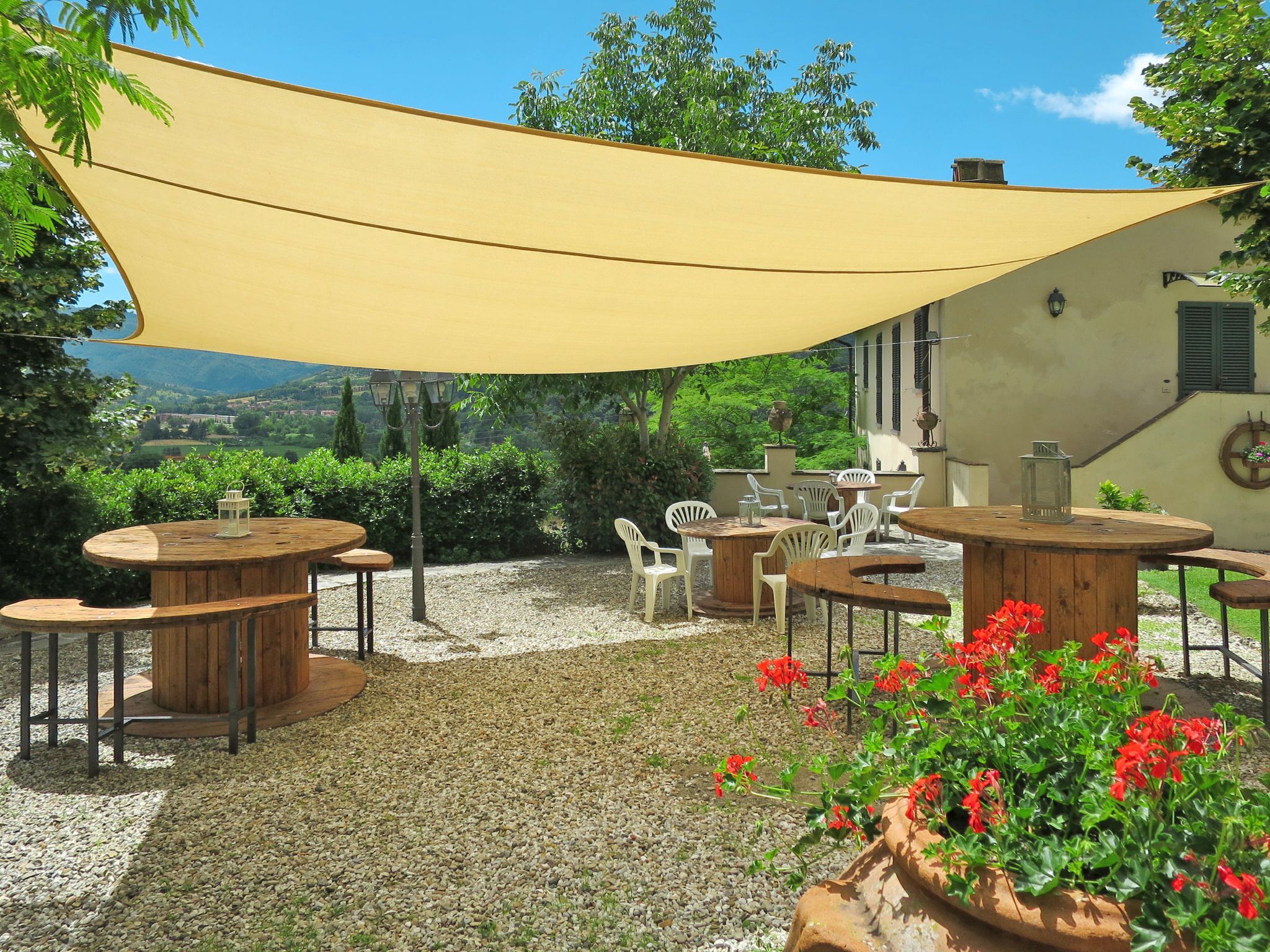 An outdoor patio with round wooden tables and chairs, a yellow shade sail, and red flowers.