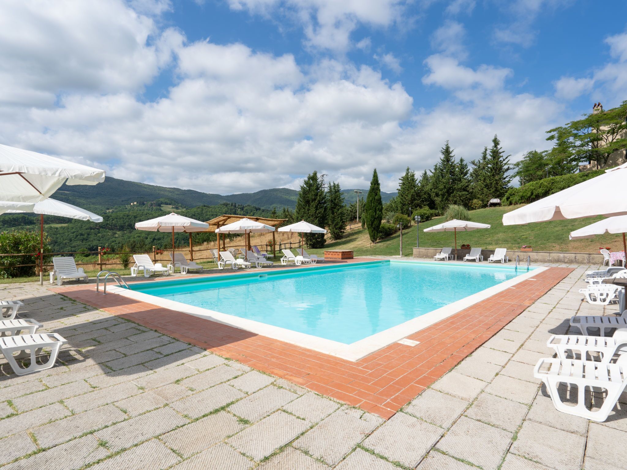 Une piscine rectangulaire entourée de chaises longues blanches et de parasols, avec en arrière-plan un paysage de montagnes et d’arbres.