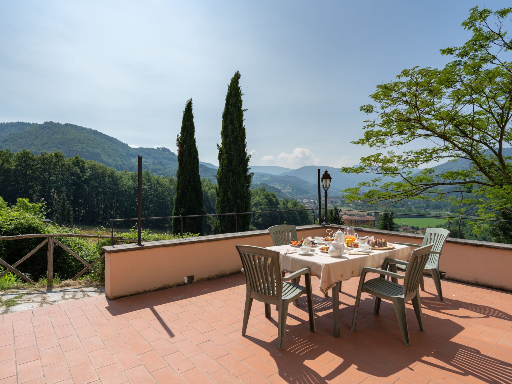 A terrace with a table set for breakfast, overlooking a scenic valley with mountains and trees.