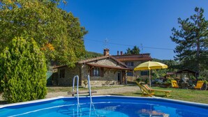 Una piscina con una casa in pietra sullo sfondo, immersa nel verde e sotto un cielo sereno.