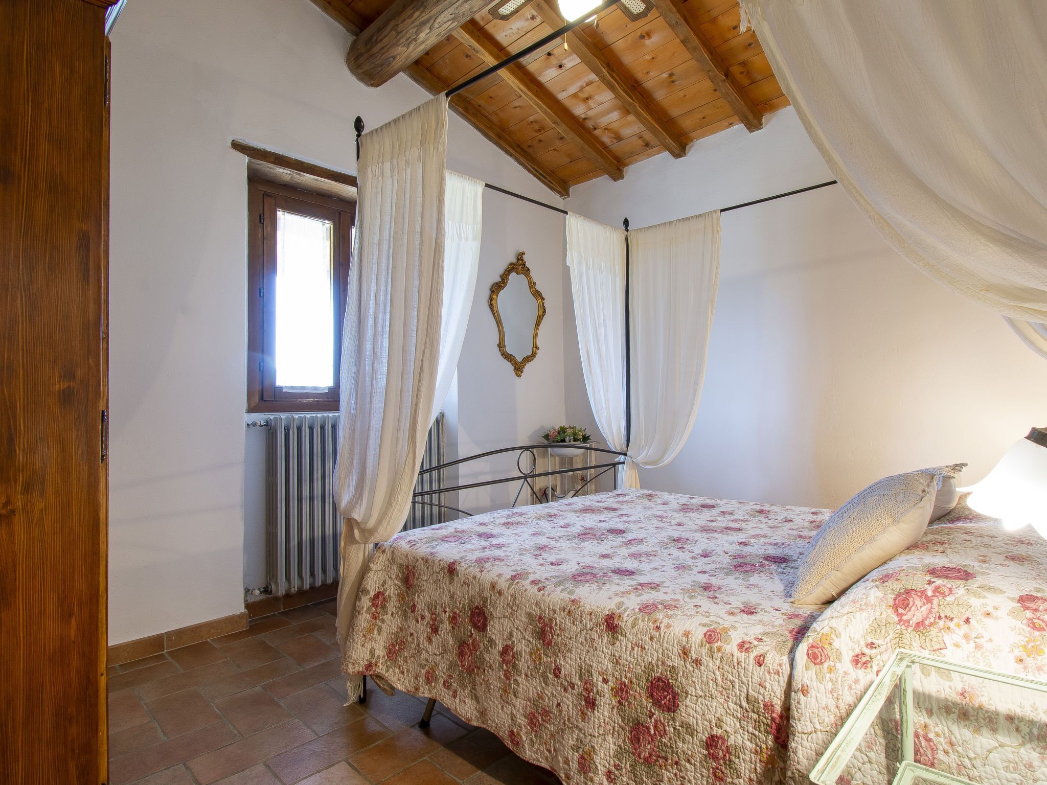Une chambre avec un plafond en bois, un lit recouvert d’une couvre-lit à motifs floraux, une fenêtre avec des rideaux et un radiateur.
