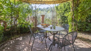 Eine überdachte Terrasse mit Tisch und Stühlen, umgeben von üppiger Bepflanzung und Blick ins Grüne.