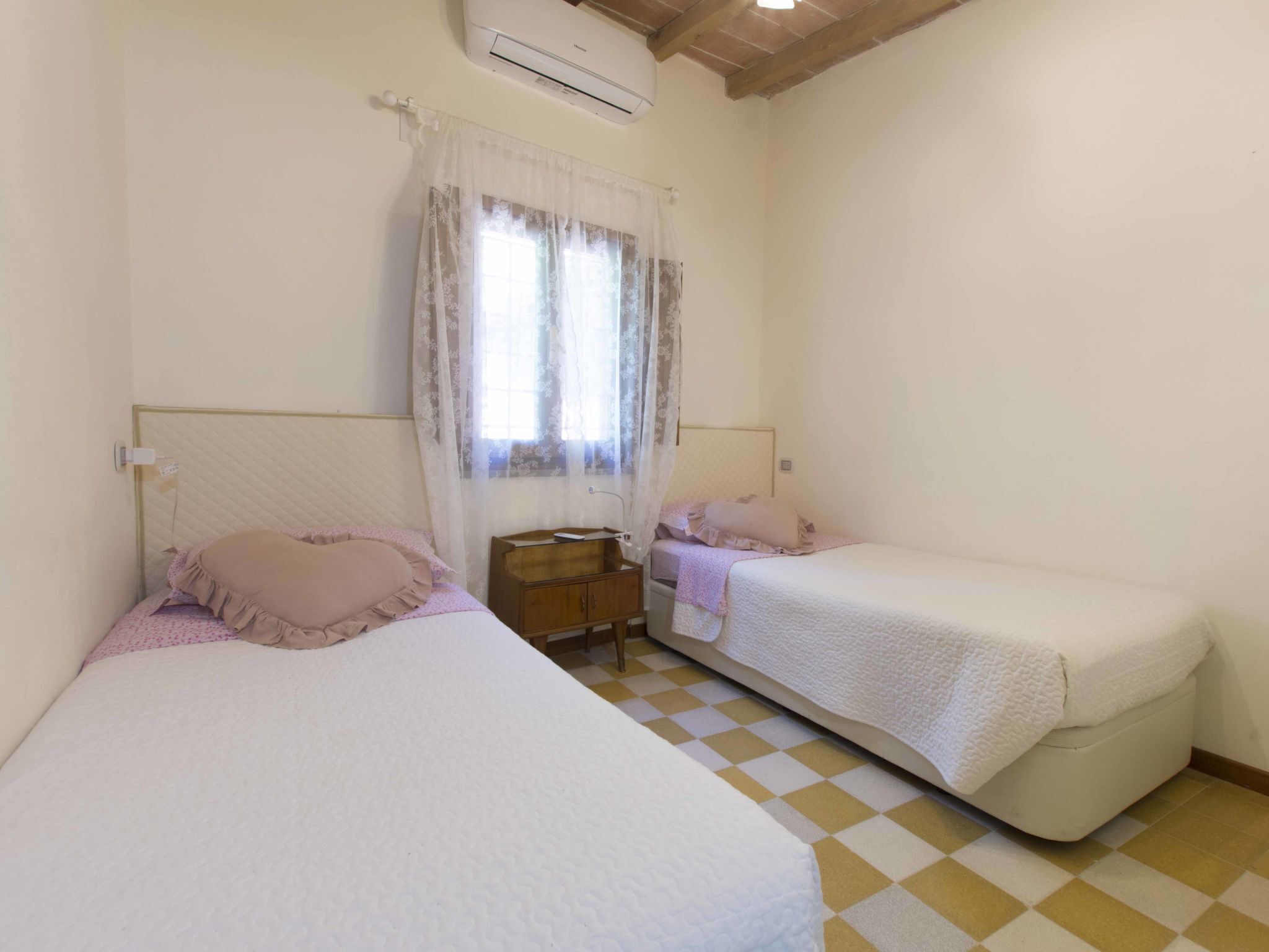 A room with two beds, a wooden nightstand, and a window with sheer curtains.