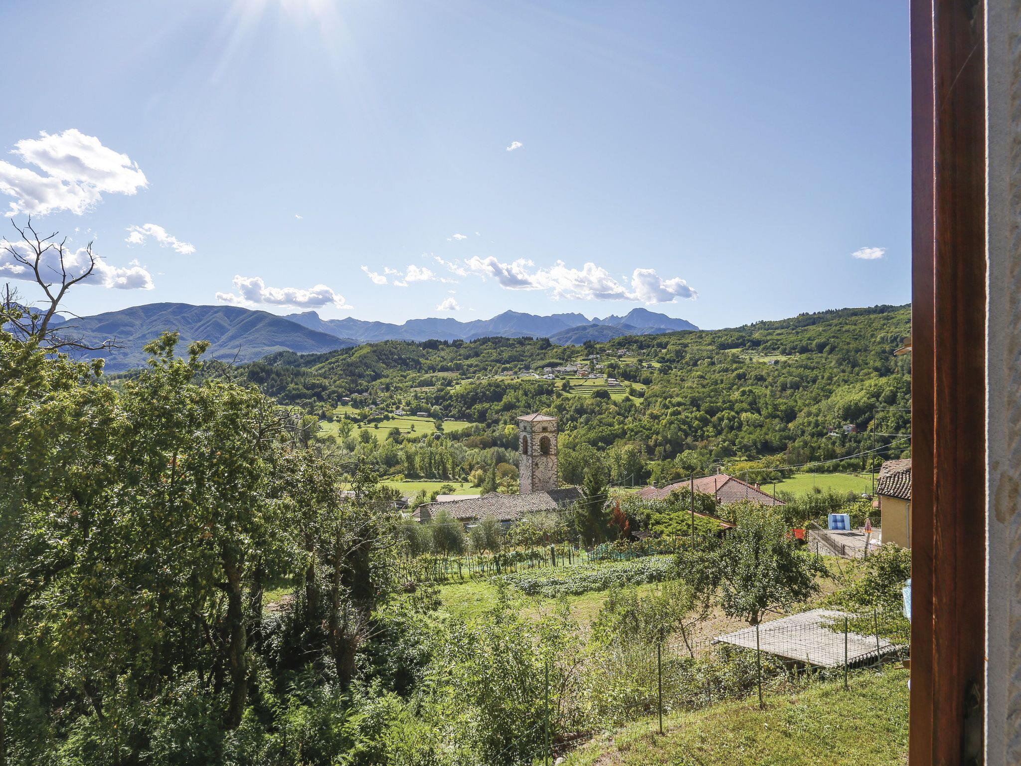 Un paysage rural avec un clocher d’église, une végétation luxuriante et des montagnes en arrière-plan.