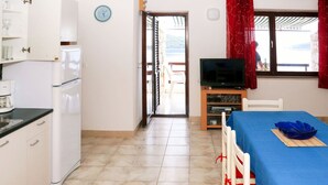 A compact room with a kitchenette, a bed with a blue bedspread, a television, and a view of the outdoors through a window.