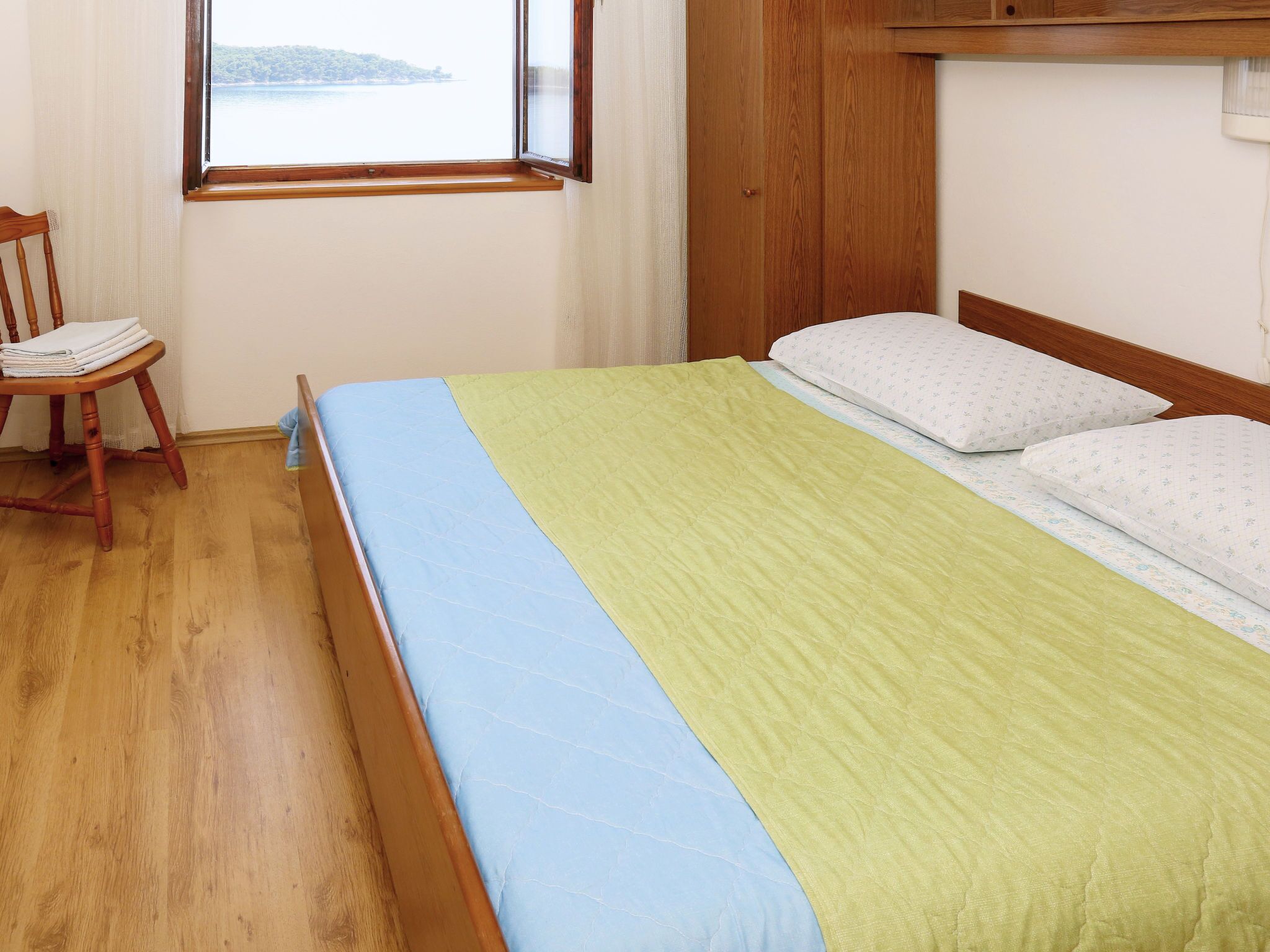 A neatly made bed with a blue and green bedspread, a wooden headboard, and a window with a view of a body of water.