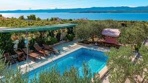 A pool area with lounge chairs and a table, surrounded by greenery and overlooking a body of water with mountains in the distance. - Vacation home Lucije by Interhome (Brač/Splitska)