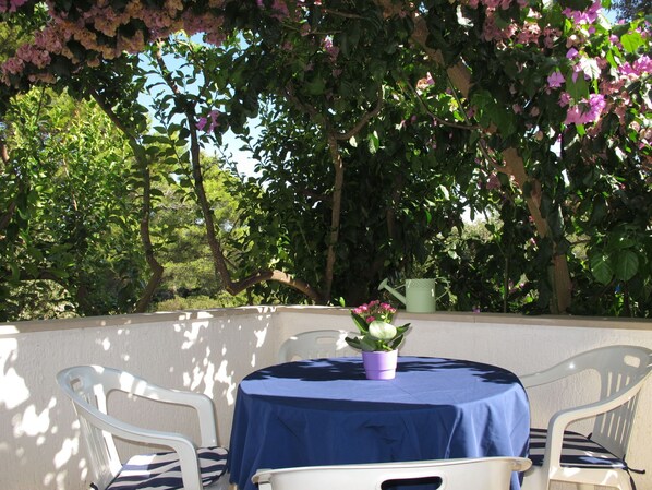A round table with a blue tablecloth, surrounded by white chairs, set against a backdrop of lush greenery and flowering vines. - Apartment Lila by Interhome (Brač/Splitska)