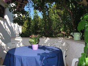 A round table with a blue tablecloth, surrounded by white chairs, set against a white wall with a potted plant and a watering can on the right. - Apartment Lila by Interhome (Brač/Splitska)