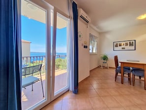 Ein Zimmer mit einer Schiebetür zu einem Balkon mit Meerblick. Es gibt einen Esstisch mit Stühlen, eine kleine Pflanze und ein gerahmtes Foto an der Wand.