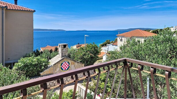 A coastal view with a clear blue sky, a stone wall with a checkered flag, and a railing in the foreground. - Apartment Culić by Interhome (Trogir)