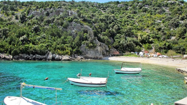 A coastal scene with boats anchored near a rocky shore, clear turquoise water, and a backdrop of dense green vegetation.