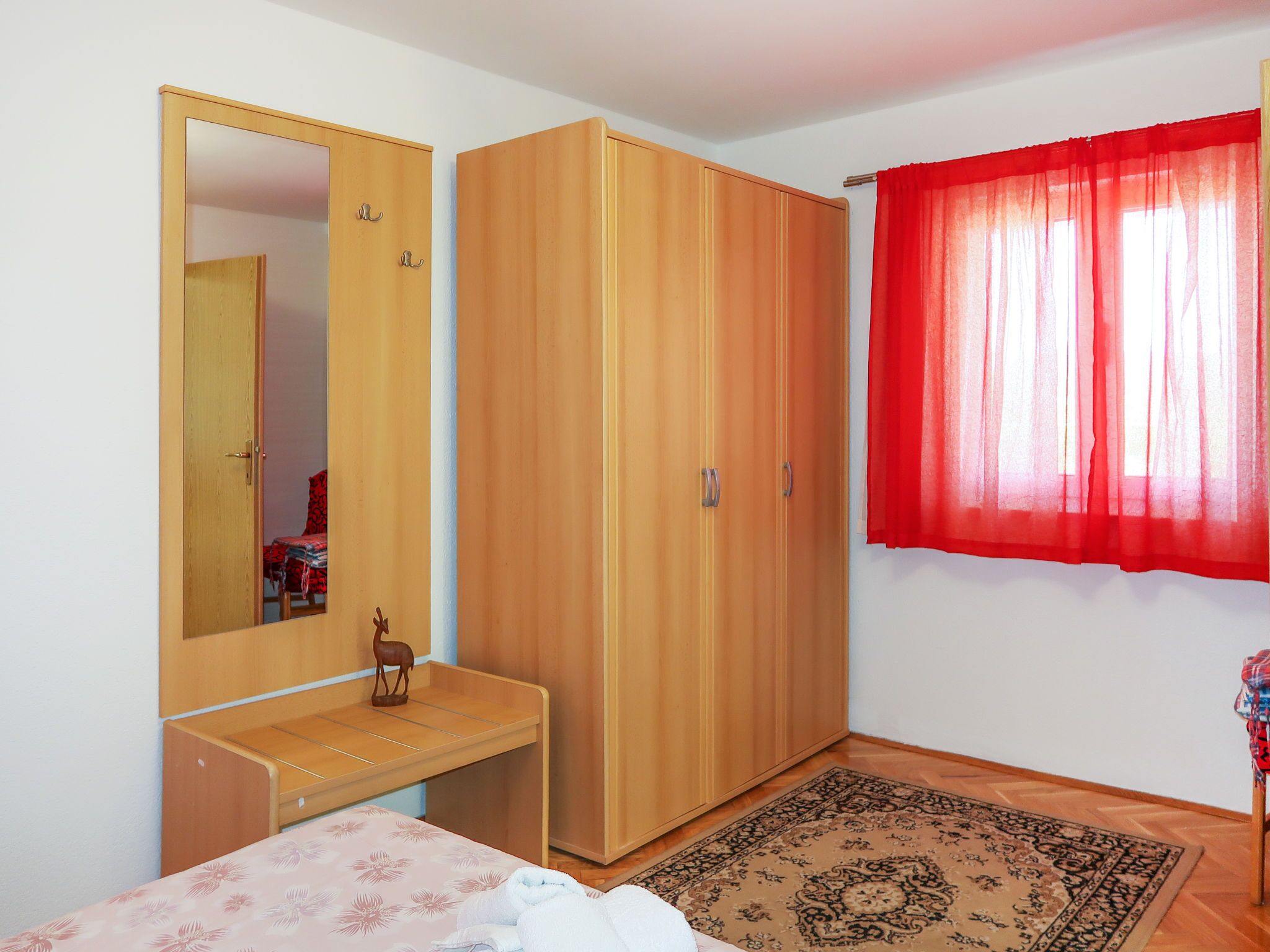 Un dormitorio con un armario de madera, una mesita de noche, un espejo, una cortina roja y una alfombra estampada.