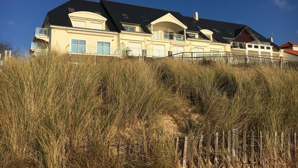 A large, multi-story building with a prominent black roof, multiple balconies, and a sandy foreground with tall grass. - Les Pieds Dans L'eau ! Accès Direct sur la Plage de Bonne Source (Pornichet)