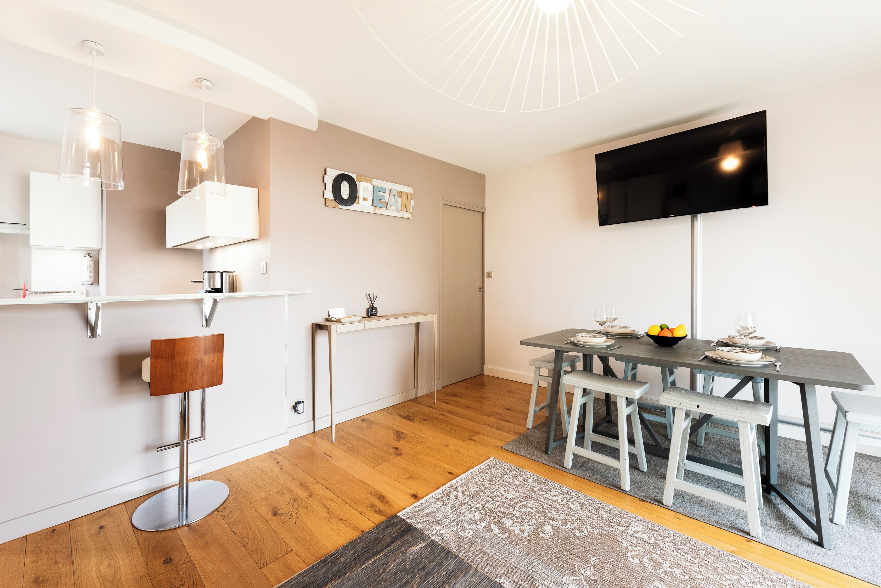 A modern living room with a dining area, a flat-screen TV mounted on the wall, and a wooden floor.