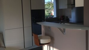 A modern kitchen with a bar stool, a built-in oven, and a window overlooking a garden. - Les Pieds Dans L'eau ! Accès Direct sur la Plage de Bonne Source (Pornichet)