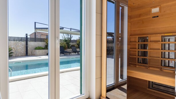 A modern room with large sliding glass doors leading to a pool area, wooden flooring, and a built-in shelving unit. - Vacation home Carmen by Interhome (Zaton (Šibenik))