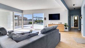 A modern living room with a grey sectional sofa, a flat-screen TV mounted on the wall, and a view of an outdoor swimming pool through large sliding doors. - Vacation home Carmen by Interhome (Zaton (Šibenik))