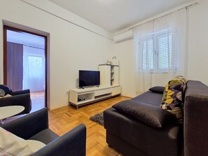 A modern living room with a flat-screen TV on a white stand, a dark sofa, a small table, and a window with blinds.