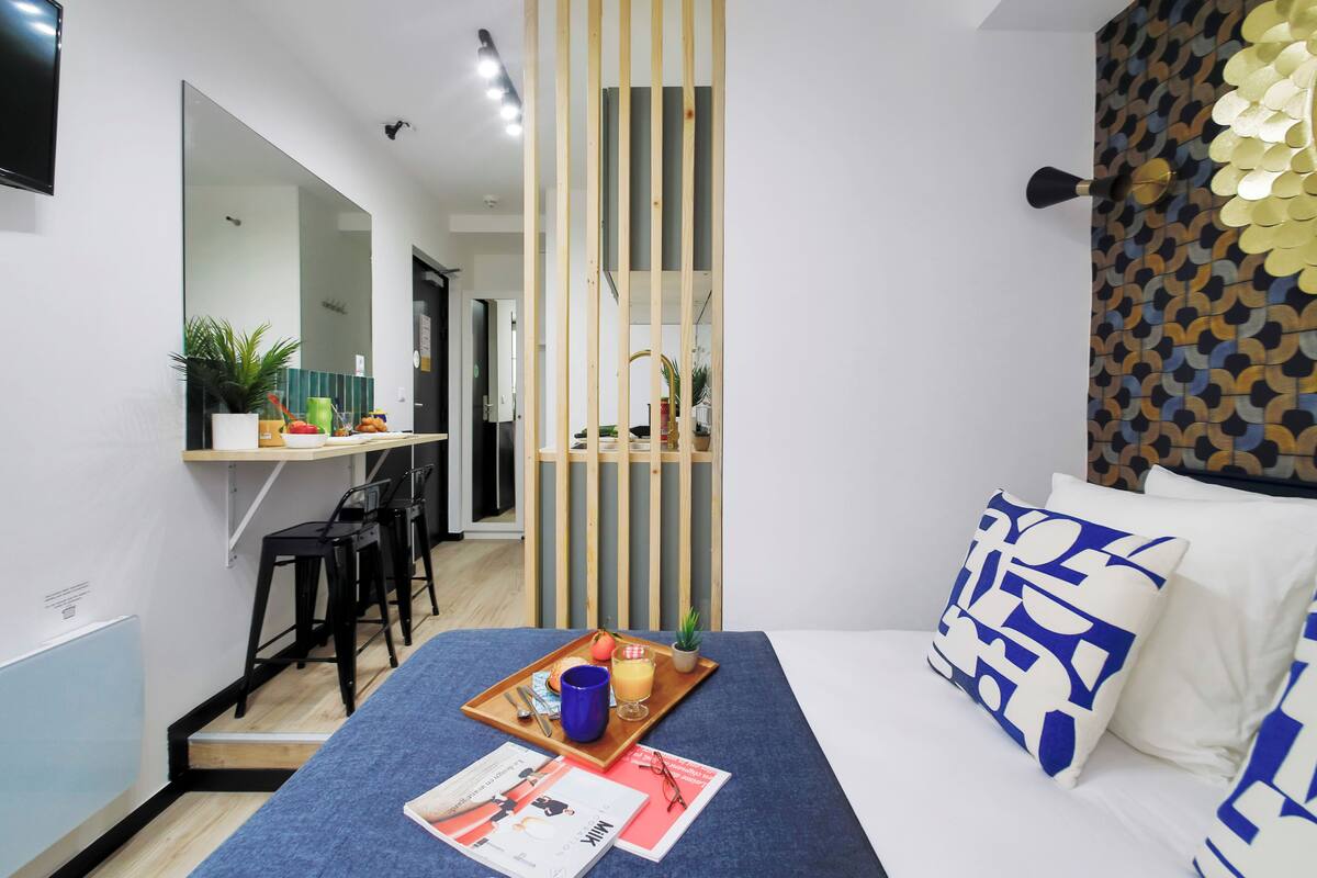 A modern hotel room with a bed, a desk, and a small dining area.