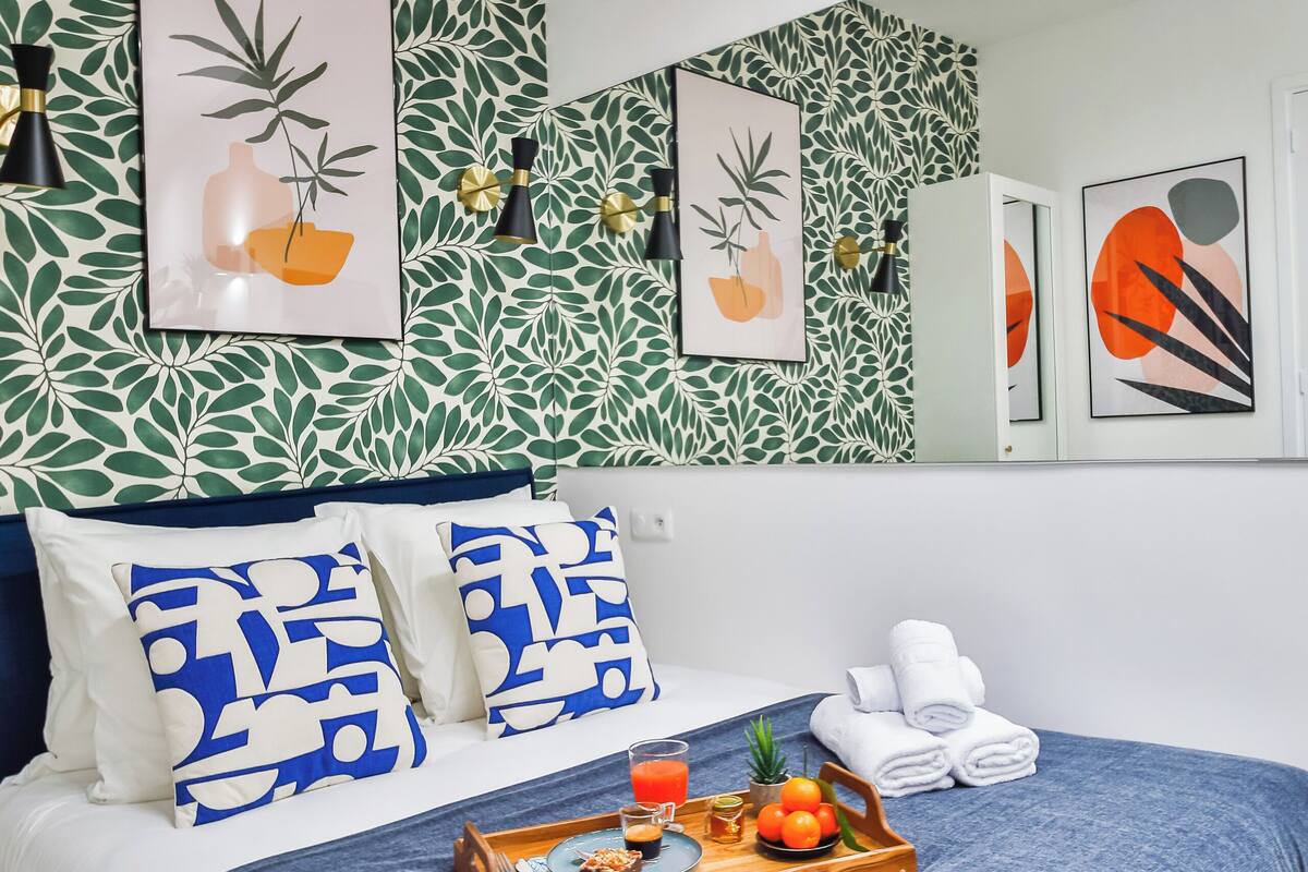 A neatly made bed with a blue bedspread, two pillows with a blue and white pattern, a tray with fruits and a glass of orange juice, and a wall with a leafy pattern and framed artwork.