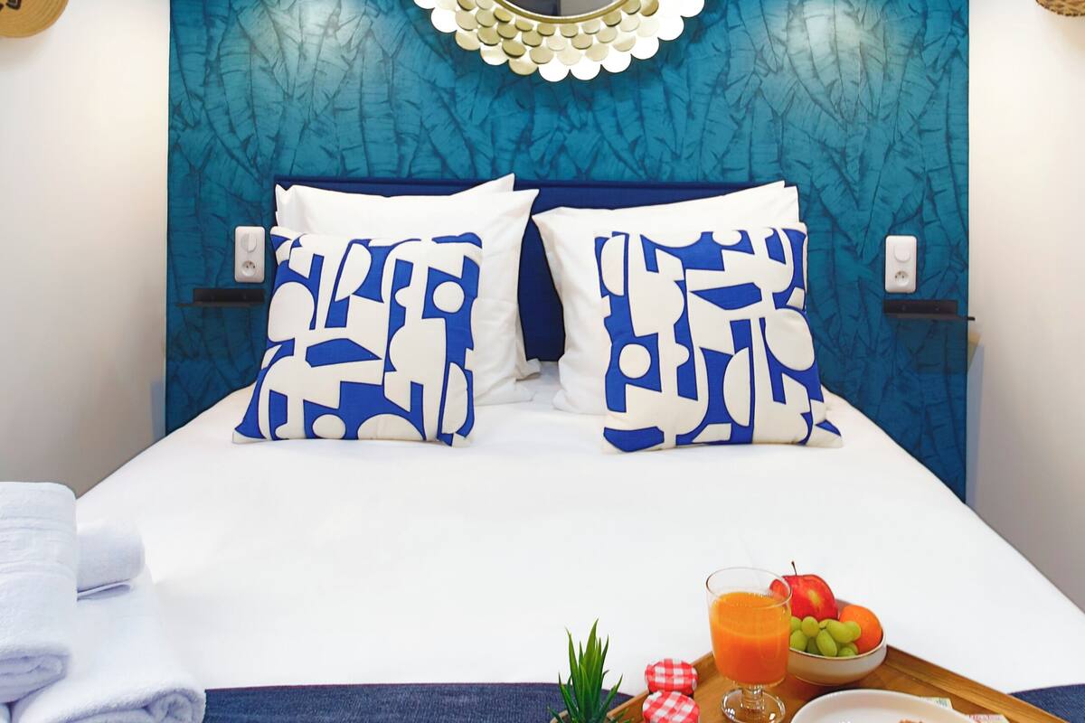 A neatly made bed with patterned pillows, a tray with breakfast including toast, fruit, and a drink, and a blue textured wall with a decorative mirror.