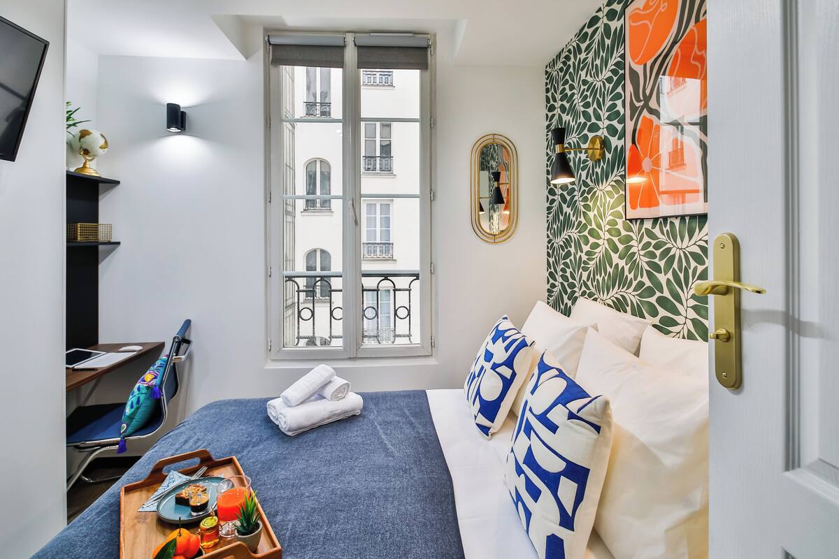 A neatly arranged hotel room with a bed, a desk, a window with a view of buildings, and a tray with breakfast items.