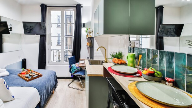 Une chambre d’hôtel de taille réduite, comprenant un lit, un petit bureau, une kitchenette et une fenêtre donnant sur le bâtiment extérieur.
