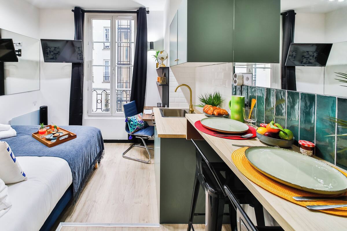 A compact hotel room with a bed, a small desk, a kitchenette, and a window with a view of the building outside.