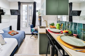 Une chambre d’hôtel de taille réduite, comprenant un lit, un petit bureau, une kitchenette et une fenêtre donnant sur le bâtiment extérieur.