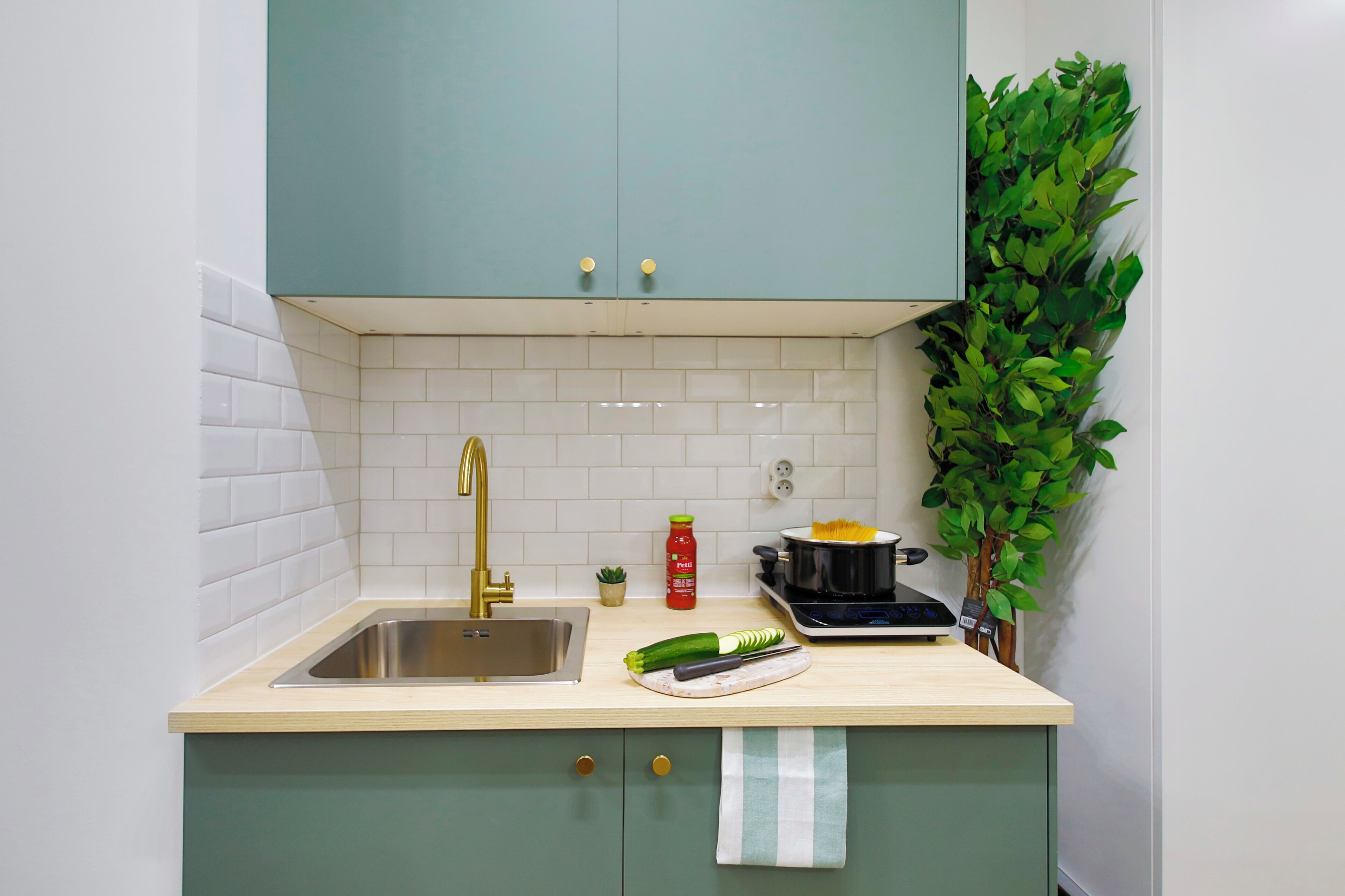 A kitchen with teal cabinets, a stainless steel sink, a small potted plant, and a cutting board with vegetables.
