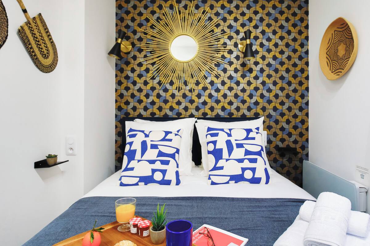 A neatly made bed with blue and white patterned pillows, a wooden tray with breakfast items, and a decorative wall with a sun-like design.