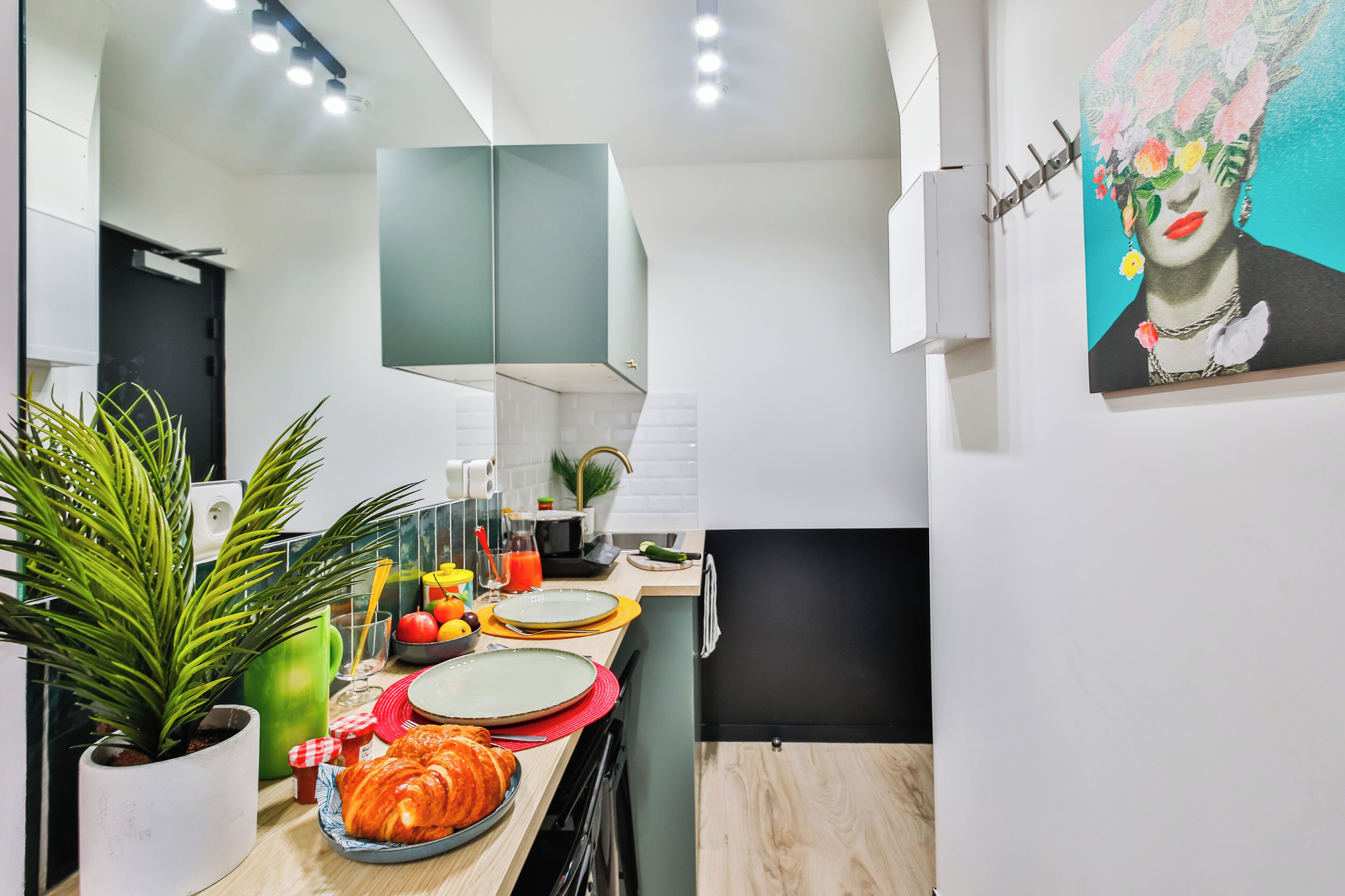 A modern kitchen with a dining area, featuring a potted plant, a fruit bowl, and a painting on the wall.