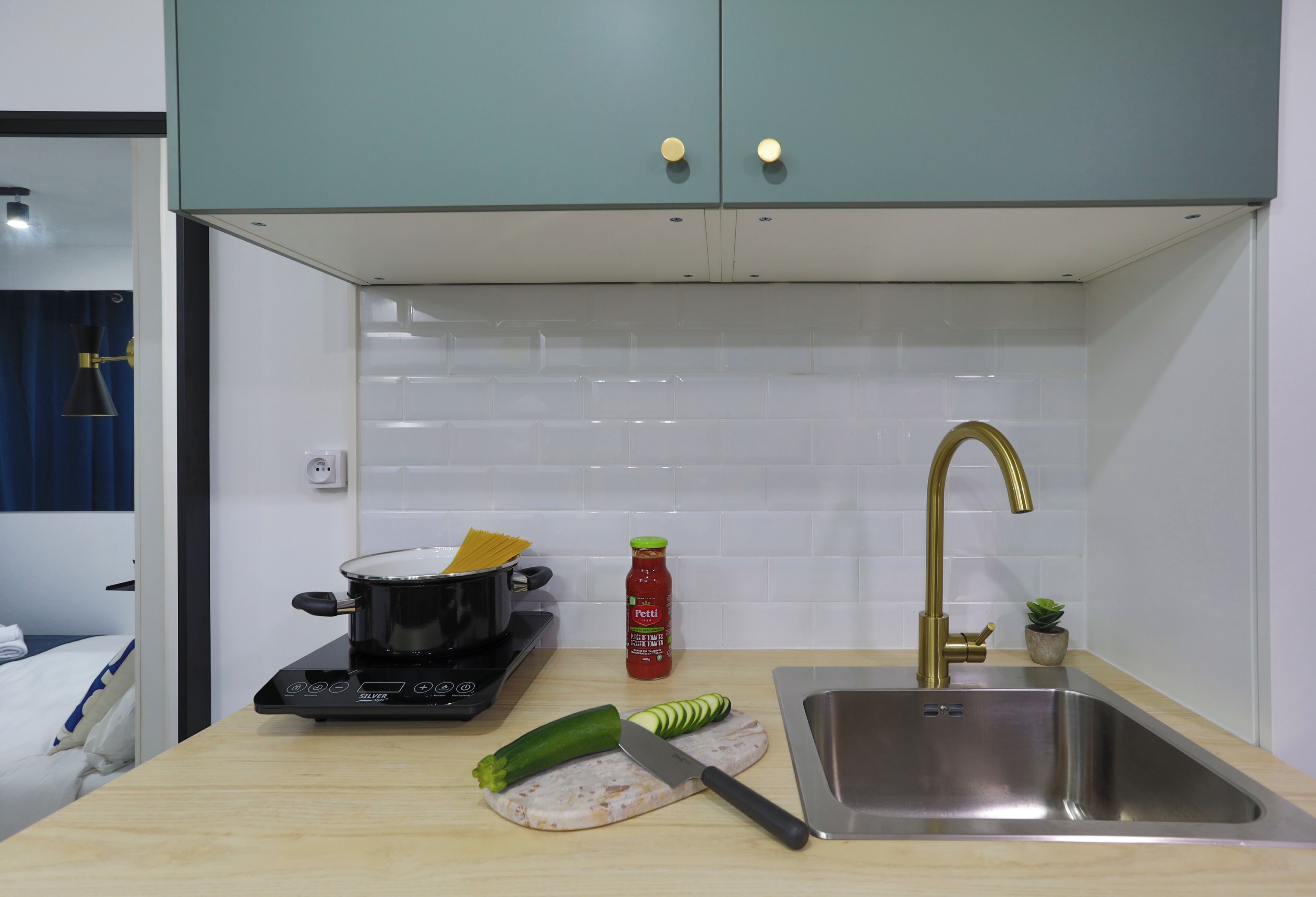 A kitchen with a stove, a sink, a cutting board with a knife, a pot, a bottle of ketchup, and a small plant.