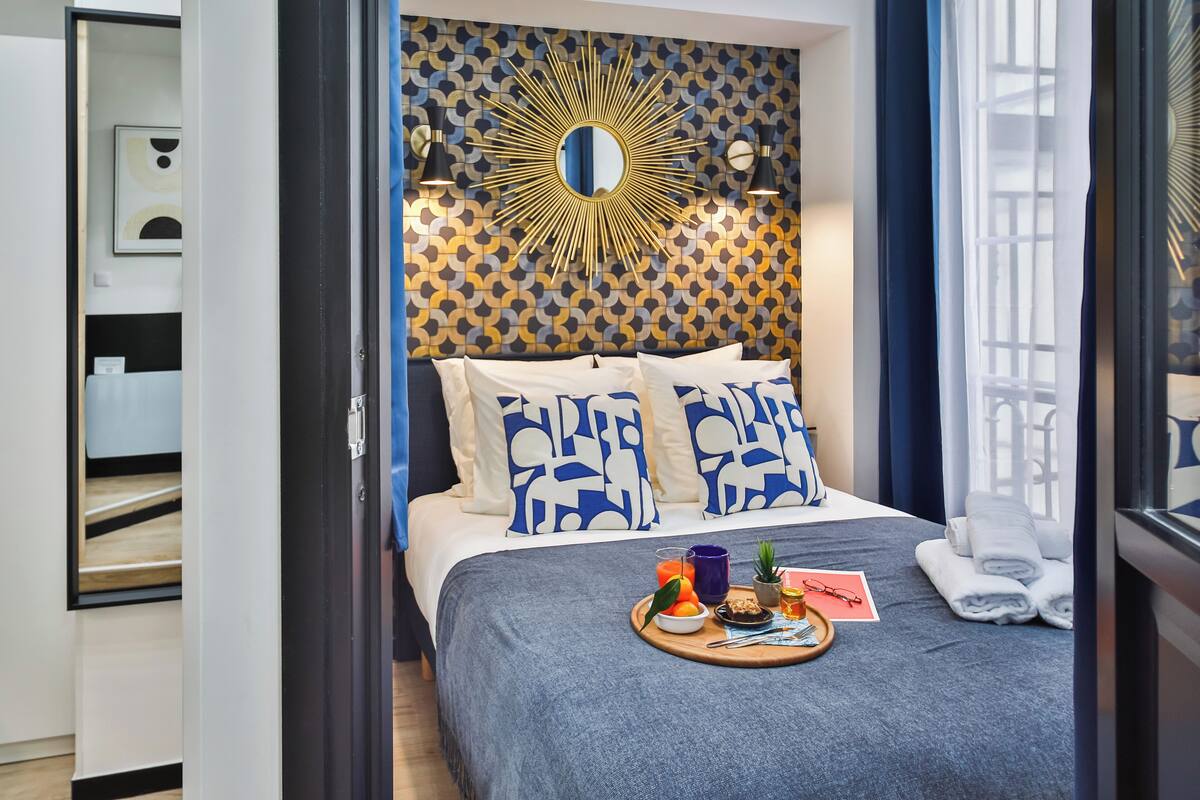 A modern hotel room with a patterned wallpaper, a large mirror, and a neatly made bed with blue and white pillows.