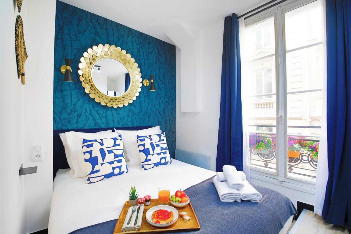 A hotel room with a bed, a breakfast tray, a window with a view of the outside, and a decorative mirror on the wall.