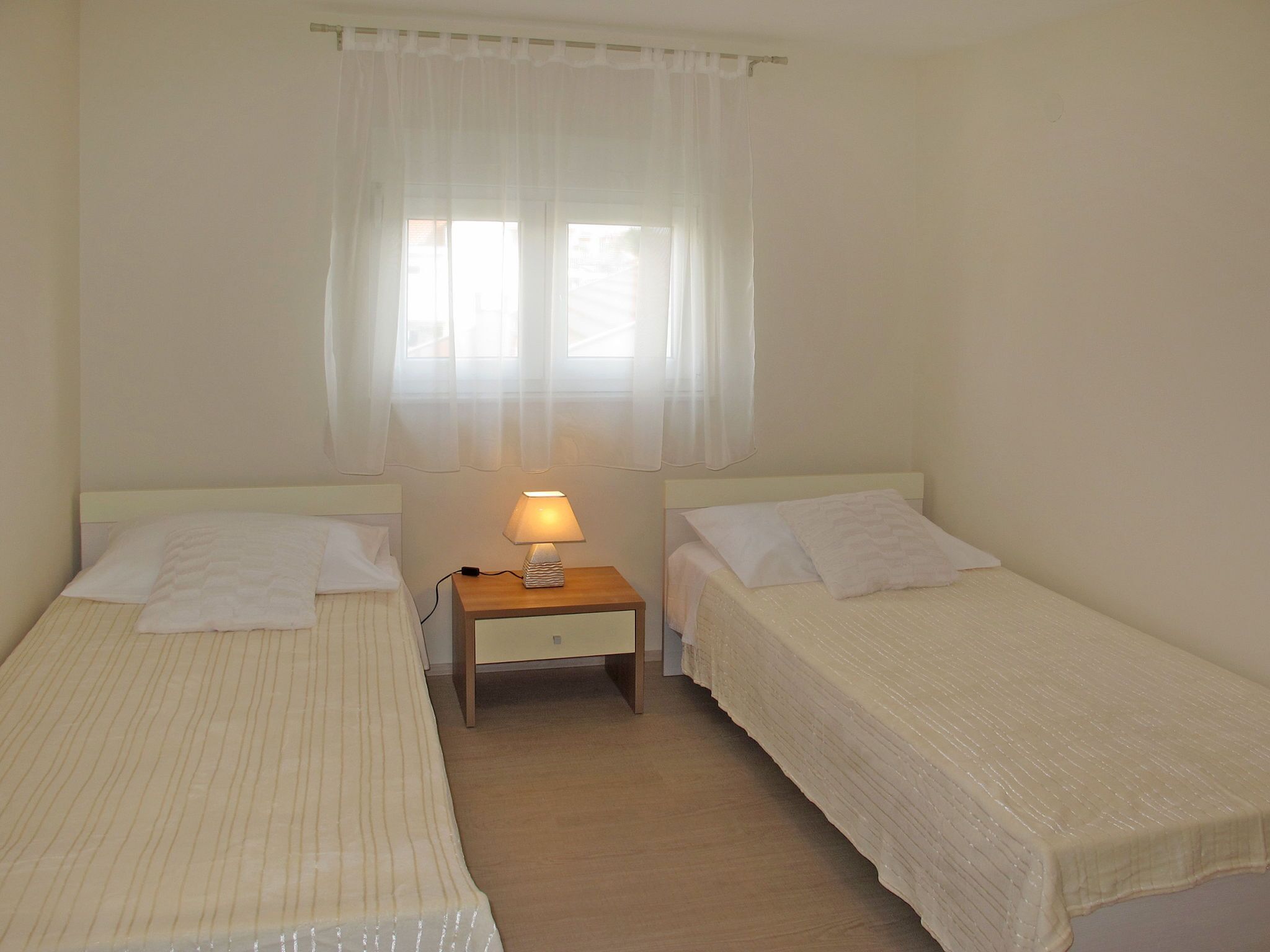 A twin room with two beds, a nightstand with a lamp, and a window with sheer curtains.